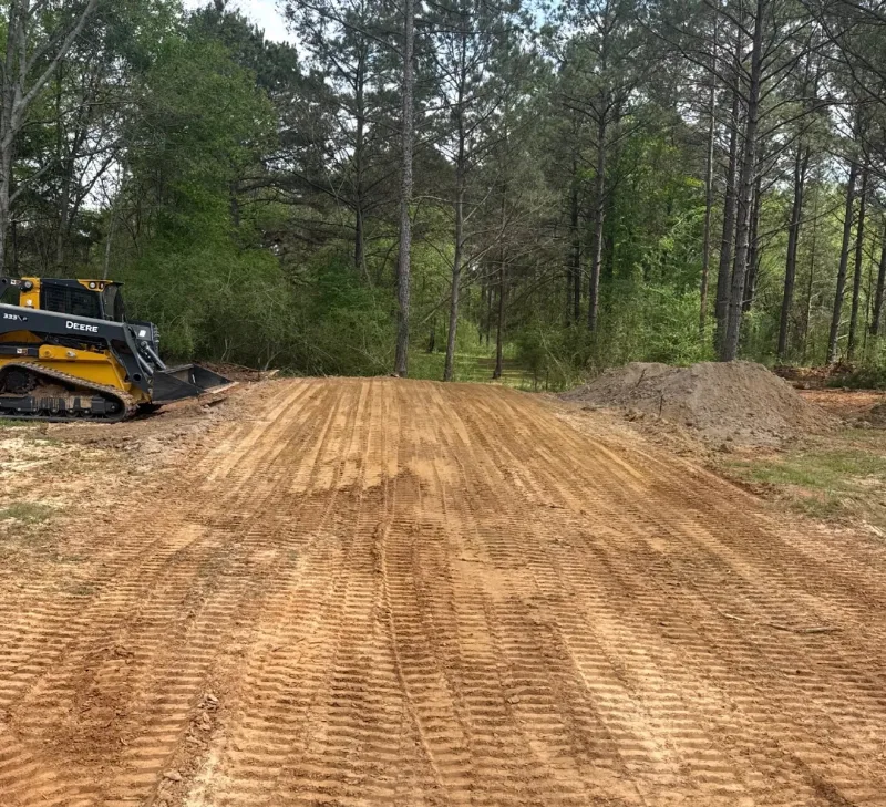 Compact track loader beside a freshly cleared and graded dirt access road running into pine woods, with a pile of spoil to one side