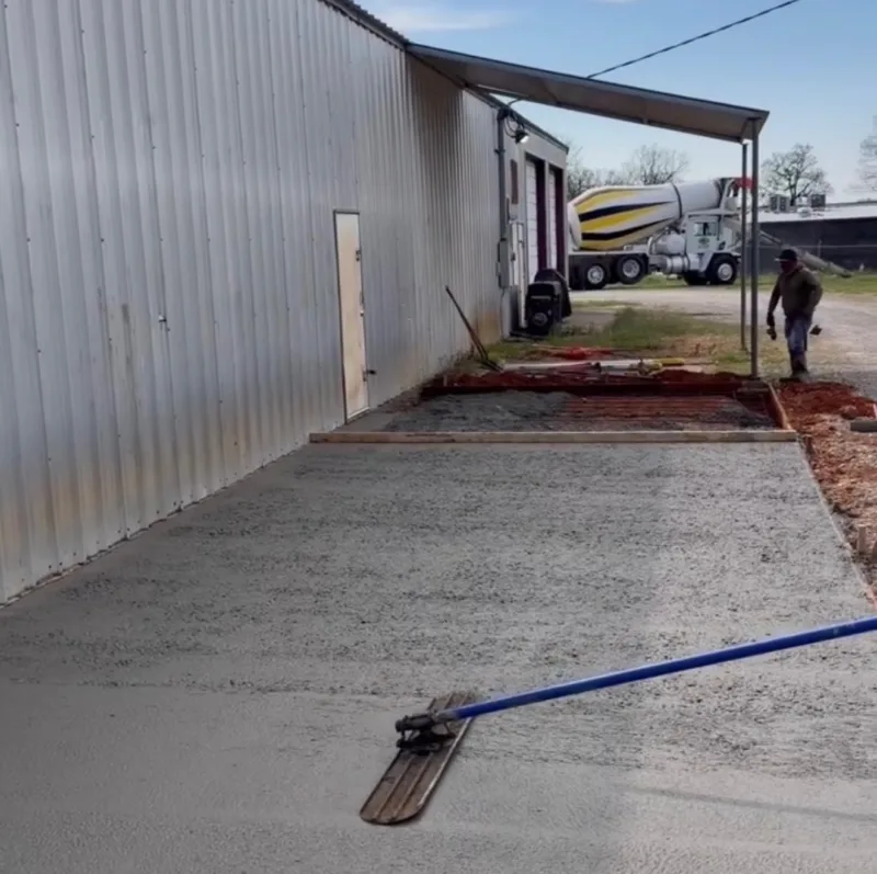 Fresh concrete apron being floated alongside a metal building, with the next formed section, a ready-mix truck and a crew member behind it