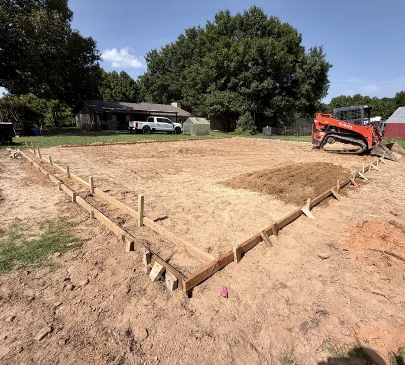 Slab forms staked out on a graded pad in a backyard, a skid steer alongside