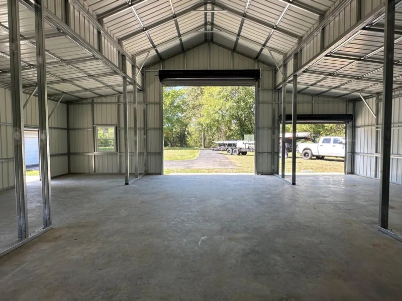 Inside a metal barn with both roll-up doors open onto a drive, trailers and a white pickup, under the steel-framed roof