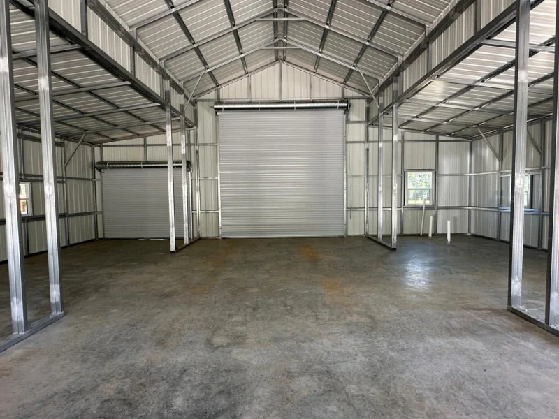 Inside a metal barn: a concrete floor, square steel framing and two white roll-up doors closed at the far end