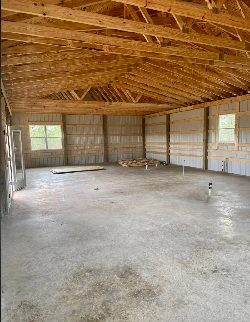 Inside a barndominium shell with wood trusses, new windows and plumbing stubbed up through the slab