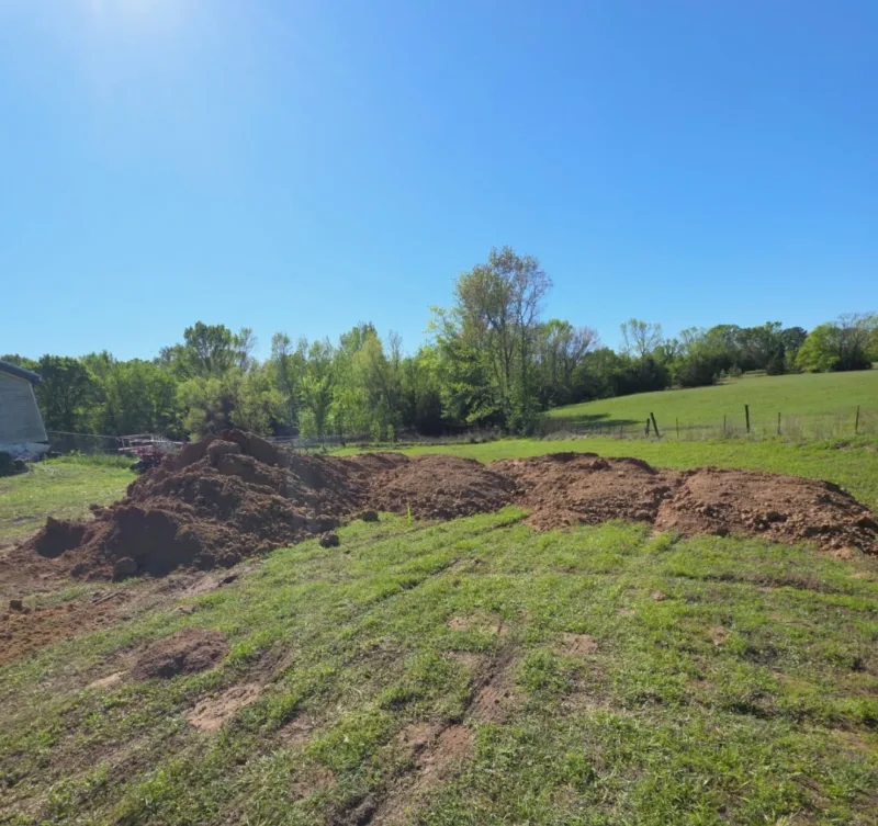Long mounds of freshly excavated dirt piled across a grassy field, with woods and a fenced pasture behind
