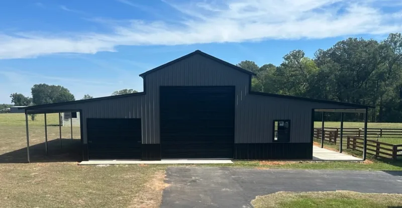 Front of a gray metal barn with a tall black roll-up door in the raised center, a second roll-up door, and an open lean-to on each side facing an asphalt drive