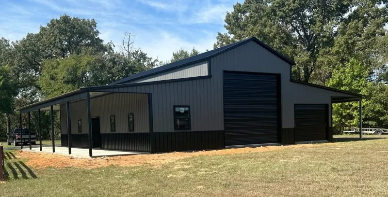 Gray metal barn with a raised center, black wainscot and roll-up doors, and open lean-tos down both sides, seen from the corner
