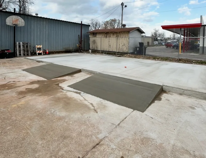 Newly poured concrete pad with two fresh ramps up from the old lot, behind a metal commercial building