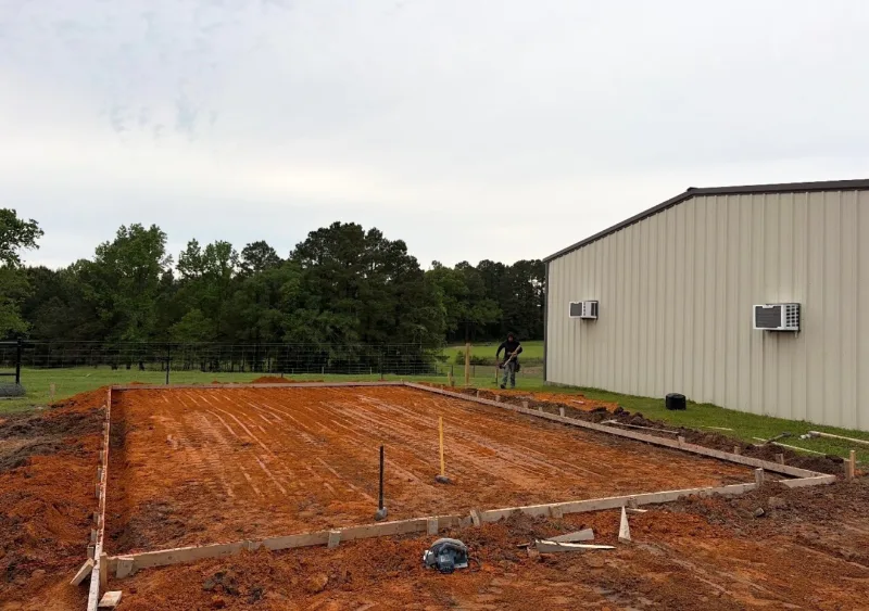 Graded red-dirt building pad with wooden forms staked around it beside a tan metal building, with a crew member at the far end