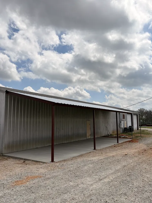 Open-sided red iron lean-to attached to a metal building under a cloudy sky
