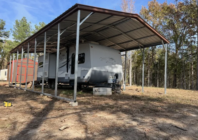 Brown metal RV cover over a travel trailer at the edge of the woods