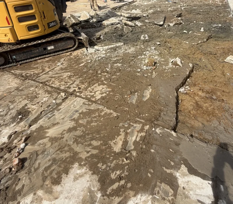 Compact excavator breaking out a saw-cut section of concrete slab, with broken concrete and wet soil exposed beside the cut