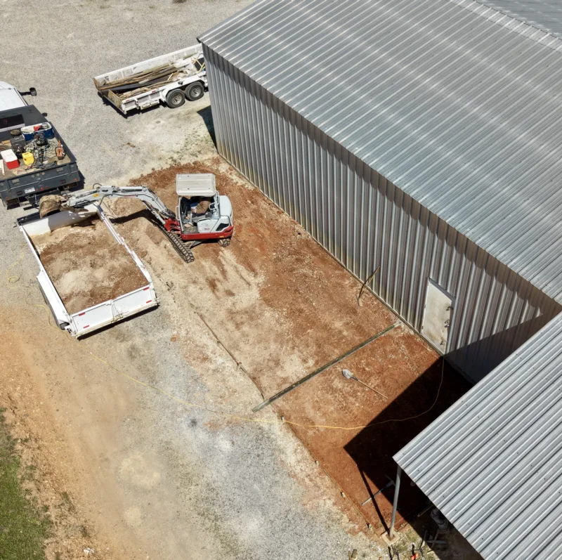 Mini excavator and trailer prepping ground beside a metal building, seen from above