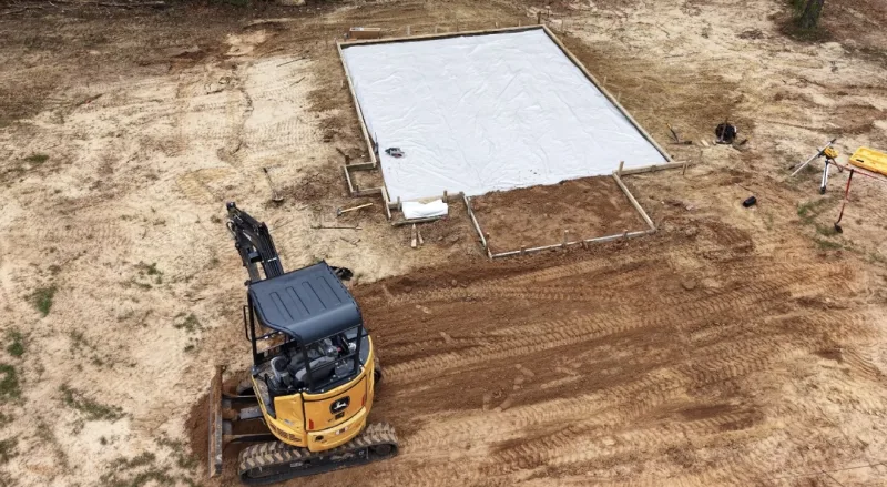 Formed slab with vapour barrier laid and an excavator grading beside it, seen from above