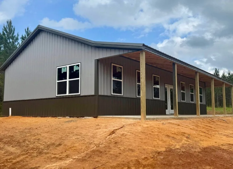 Grey and brown barndominium with a long covered porch on a cleared lot in the pines